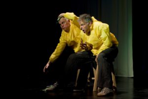 Zwei Schauspieler in gelben Regenponchos stehen gebückt und scheinbar gegen Wind kämpfend auf der Bühne.