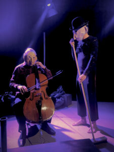 Ein Mann mit Maske sitzt auf einem Stuhl und spielt Cello. Ein zweiter Mann mit Maske steht fegend rechts neben ihm. Der Bildraum ist scheinbar eine Bühne und in violettem Licht ausgeleuchtet.