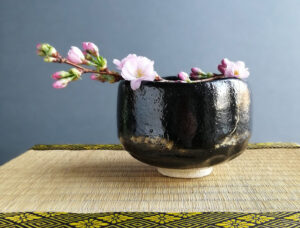 Eine schwarze japanische Teeschale mit einem Kirschblütenast steht vor einem grauen Hintergrund auf einem Tisch mit einer Rattanunterlage.