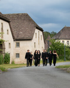 Sechs Männer und Frauen in schwarzen Kleidern gehen auf einer geteerten Straße, die eine Kurve macht. Häuser stehen entlang dieser ländlichen Straße.